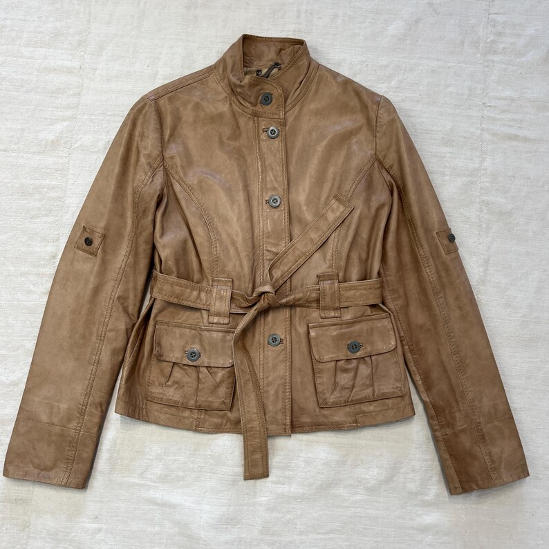 K&ouml;nnte beinhalten: Eine braune Lederjacke mit G&uuml;rtel und Taschen. Die Jacke hat einen Stehkragen und einen Knopfverschluss.
