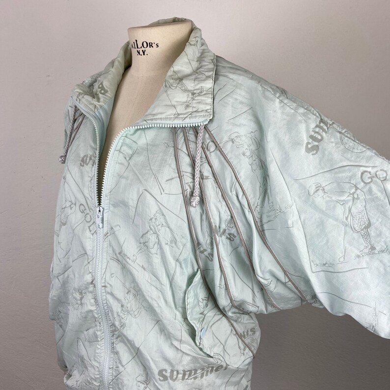 May include: A light blue vintage windbreaker jacket with a full zipper and a hood. The jacket features a golf-themed pattern with the words "Summer Golf" printed on it. The jacket is displayed on a mannequin.
