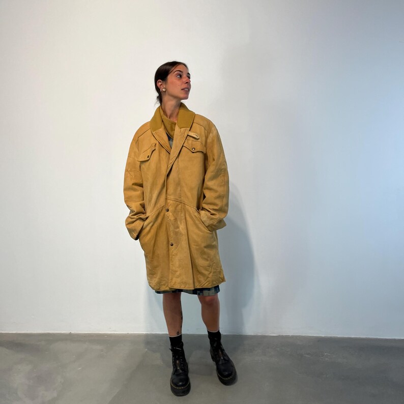May include: A long, tan suede coat with a button closure and a collar. The coat has two large pockets on the front and a small pocket on the chest. The coat is worn by a person with dark hair and is standing on a concrete floor.