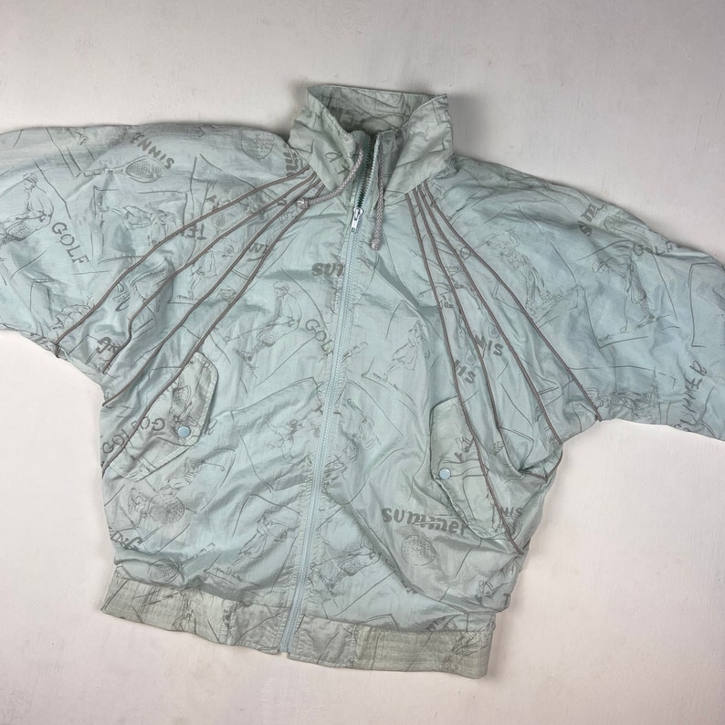 May include: Light blue vintage windbreaker jacket with a full zipper, featuring a pattern of golf and tennis figures. The jacket has the word "SUMMER" printed on it.