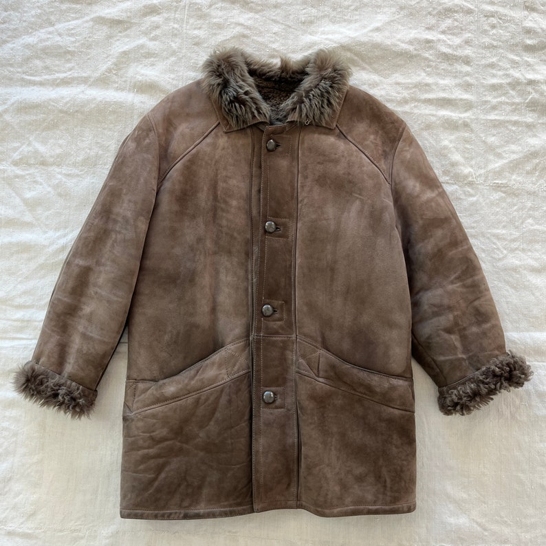 Puede incluir: Una chaqueta de piel de oveja marr&oacute;n de ante con cuello y pu&ntilde;os de piel. La chaqueta tiene cierre de botones y dos bolsillos de solapa.