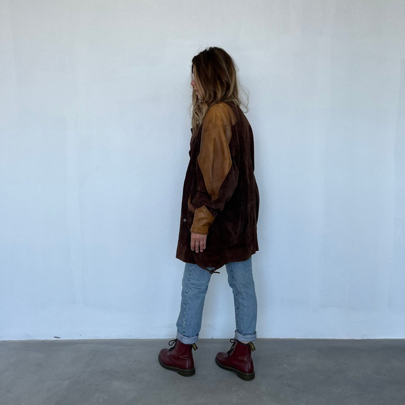 May include: A brown suede jacket with brown leather sleeves. The jacket has a zipper closure and a buttoned flap pocket on the left side. The model is wearing blue jeans and maroon Doc Martens.