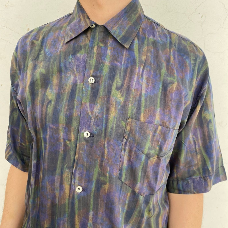 May include: A short-sleeved button-down shirt with a colorful abstract print in shades of blue, green, and purple. The shirt has a pocket on the left side.