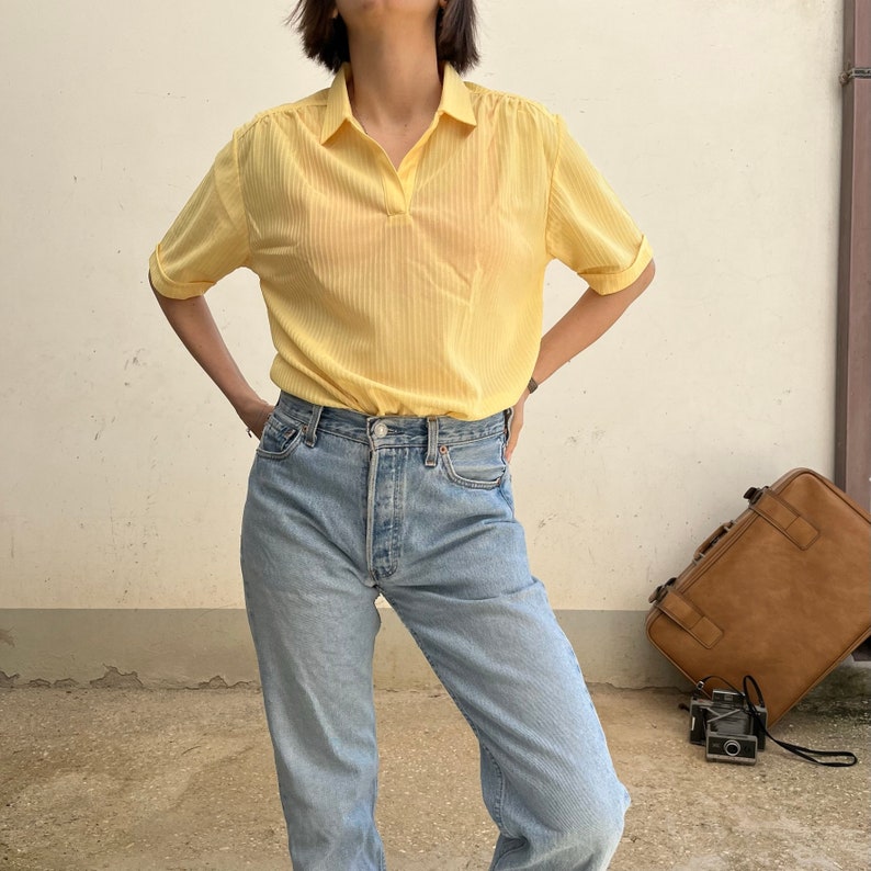 May include: A yellow striped collared shirt with short sleeves. The shirt is tucked into a pair of light blue jeans.