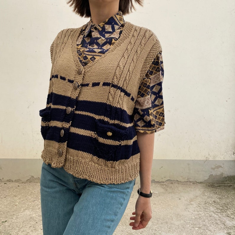 K&ouml;nnte beinhalten: Eine beige-marineblaue Strickweste mit V-Ausschnitt, Knopfverschluss und zwei Vordertaschen. Die Weste hat ein gestreiftes Design und wird &uuml;ber einem gemusterten Hemd und einer blauen Jeans getragen. Das Outfit wird durch eine schwarze Uhr erg&auml;nzt.