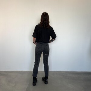 May include: A person wearing a black cropped t-shirt and dark gray jeans with a ripped design on the back of the legs. The person is also wearing black boots. The background is a plain white wall.