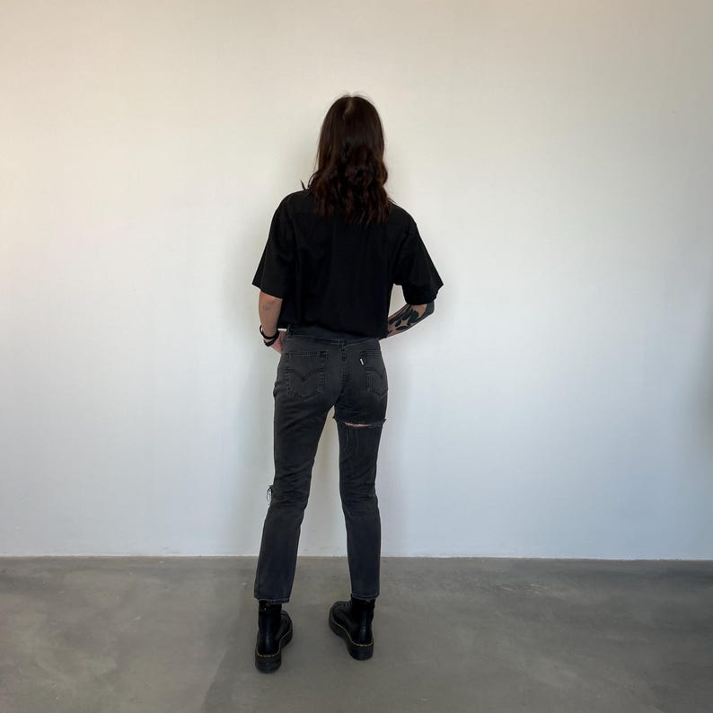 May include: A person wearing a black cropped t-shirt and dark gray jeans with a ripped design on the back of the legs. The person is also wearing black boots. The background is a plain white wall.