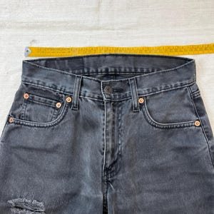 May include: Close-up of a pair of faded black denim jeans. The jeans have a button closure, classic five-pocket design, and copper rivets. A yellow measuring tape is placed across the waistband, showing measurements in inches.