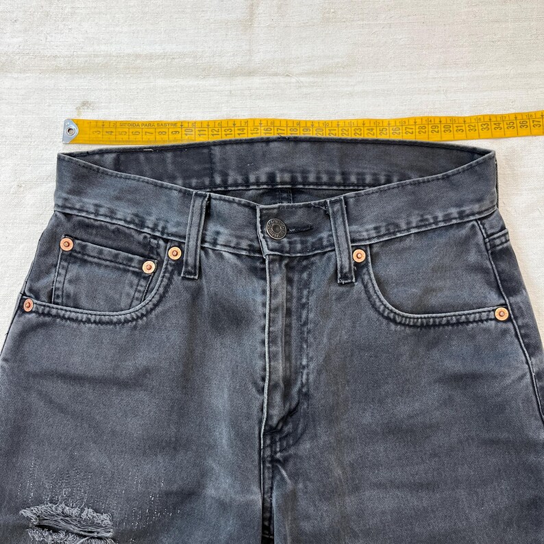 May include: Close-up of a pair of faded black denim jeans. The jeans have a button closure, classic five-pocket design, and copper rivets. A yellow measuring tape is placed across the waistband, showing measurements in inches.