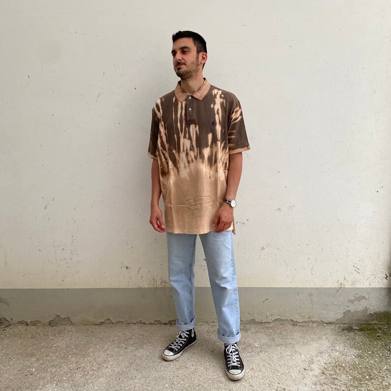 K&ouml;nnte beinhalten: Ein braunes und beigefarbenes Polohemd mit Krawattenmuster und einer Knopfleiste mit drei Kn&ouml;pfen. Das Hemd wird mit hellblauen Jeans und schwarzen High-Top-Sneakern kombiniert. Eine silberne Uhr ist am Handgelenk zu sehen.