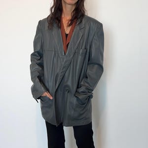 Op de afbeelding: Een grijs leren blazer met een dubbele rij knopen en een lange, ontspannen pasvorm.