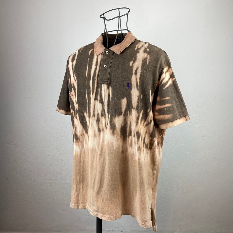 K&ouml;nnte beinhalten: Ein braunes Poloshirt mit gebleichtem Batik-Effekt. Das Shirt hat einen hellbraunen Kragen und ein kleines lila Logo auf der Brust. Das Shirt wird auf einer schwarzen Schaufensterpuppe pr&auml;sentiert.