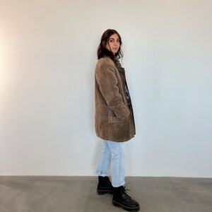 Puede incluir: Una chaqueta de ante marr&oacute;n con cuello de piel y bolsillos. La chaqueta la lleva puesta una persona con el pelo largo y casta&ntilde;o y est&aacute; de pie frente a una pared blanca.