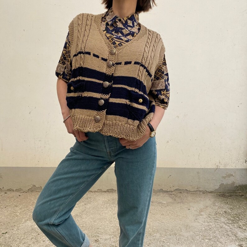 K&ouml;nnte beinhalten: Ein beigefarbener und marineblauer Strickcardigan mit kurzen &Auml;rmeln und goldenen Kn&ouml;pfen. Der Cardigan wird &uuml;ber einer gemusterten Bluse getragen und mit hellblauen Jeans kombiniert. Das Outfit ist im Vintage-Stil.