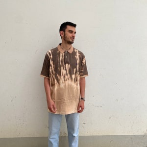 K&ouml;nnte beinhalten: Braunes und beiges Batik-Poloshirt mit lila Logo auf der Brust. Das Shirt hat einen klassischen Kragen und einen Knopfverschluss. Das kurz&auml;rmlige Shirt wird mit hellblauen Jeans getragen.