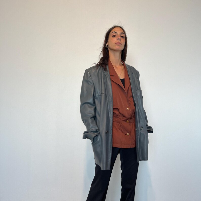 Op de afbeelding: Een vrouw draagt ​​een donkergrijze leren blazer over een roestkleurig overhemd met knopen en een getailleerde taille. Ze draagt ​​een zwarte broek.