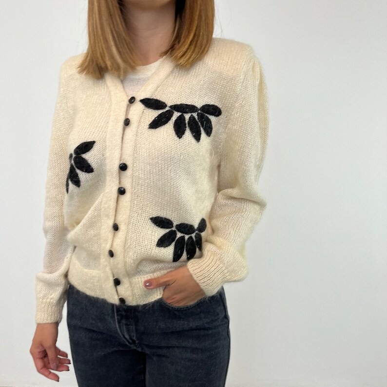 May include: A cream-coloured cardigan with black embroidered leaf designs. The cardigan has a button-up front and long sleeves.
