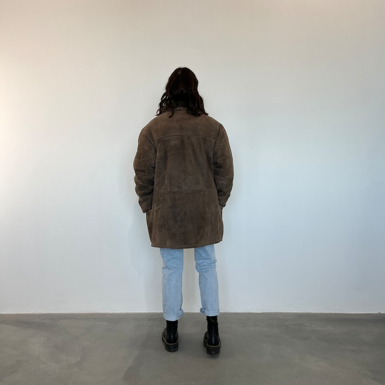 Puede incluir: Una chaqueta de ante marr&oacute;n de corte largo y recto. La chaqueta tiene cuello y cierre de botones. La persona que lleva la chaqueta est&aacute; de pie frente a una pared blanca.