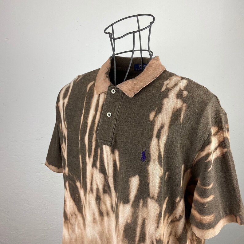 K&ouml;nnte beinhalten: Ein braunes Poloshirt mit einem Bleichmittel-Design. Das Shirt hat einen hellbraunen Kragen und kurze &Auml;rmel. Es hat ein kleines, lila Logo auf der Brust und drei wei&szlig;e Kn&ouml;pfe.
