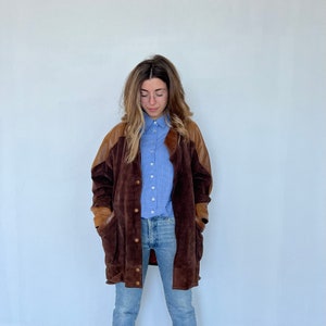 May include: A brown suede jacket with leather accents. The jacket has a button-up front and two large pockets. The model is wearing a light blue plaid shirt and blue jeans.