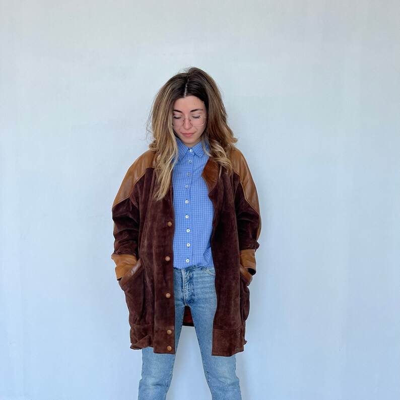 May include: A brown suede jacket with leather accents. The jacket has a button-up front and two large pockets. The model is wearing a light blue plaid shirt and blue jeans.