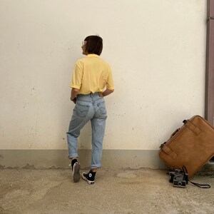 May include: A person wearing a yellow collared shirt, blue jeans, and black sneakers stands in front of a white wall. A brown suitcase and a camera are on the ground.