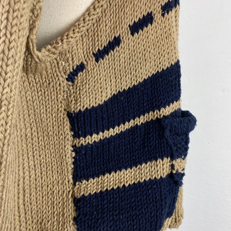 K&ouml;nnte beinhalten: Nahaufnahme eines gestrickten Kleidungsst&uuml;cks in Beige und Marineblau. Das Kleidungsst&uuml;ck hat eine beige Basis mit diagonalen marineblauen Streifen. Das Strickwerk hat ein strukturiertes Aussehen mit sichtbaren Maschen und einem Taschen Detail.