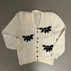 May include: A cream knitted cardigan with black velvet floral embellishments. The cardigan has a V-neck and button closure.