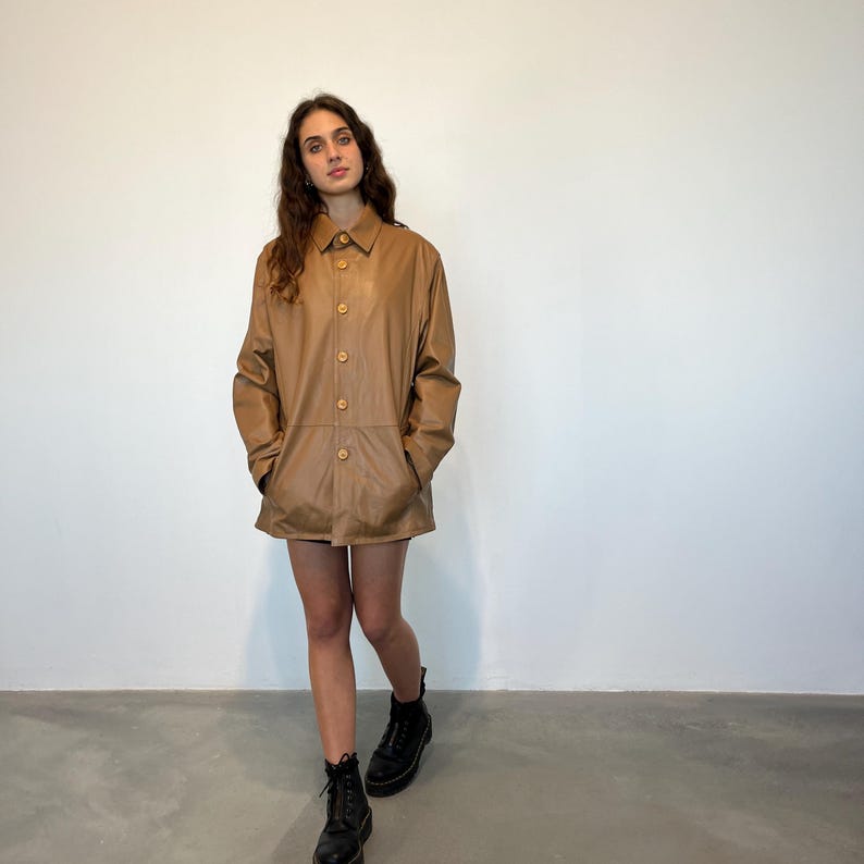 May include: A brown leather jacket with a button-up front and long sleeves. The jacket is worn by a person with long brown hair and is standing on a light brown floor.
