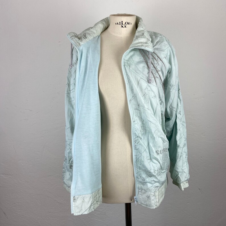 May include: A light blue jacket with a full zipper, displayed on a mannequin. The jacket has a hood and a light blue interior. The exterior features a subtle, patterned design in a lighter shade of blue. The jacket is open, revealing the interior.