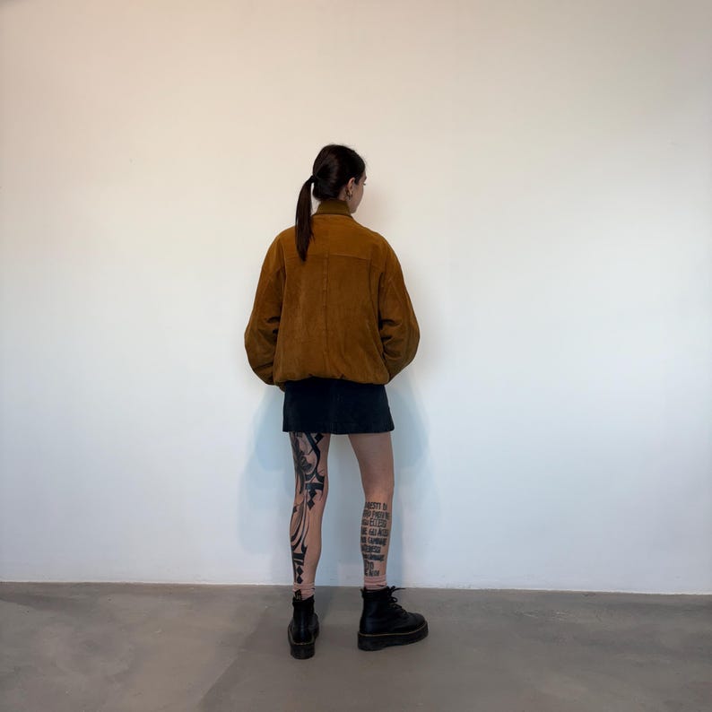 Op de afbeelding: Een bruine corduroy jas, zwarte rok en zwarte laarzen. De jas heeft een kraag en een knoopsluiting. De outfit wordt gedragen door een persoon met been tatoeages.