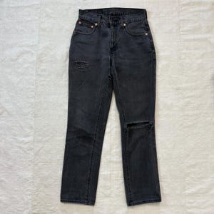 May include: Dark gray denim jeans with a classic five-pocket design. The jeans feature distressed details, including a ripped knee and a worn patch on the thigh. The jeans have a straight leg cut and a button and zipper closure.