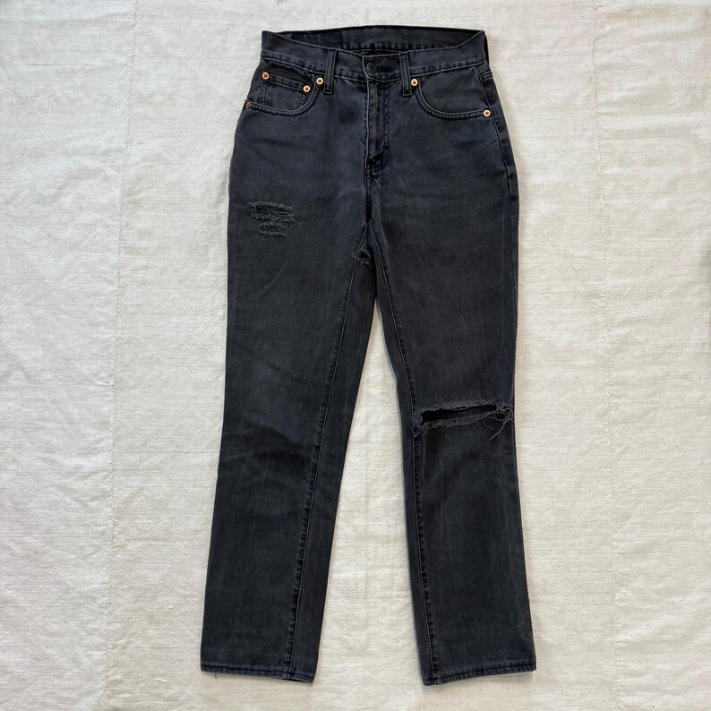 May include: Dark gray denim jeans with a classic five-pocket design. The jeans feature distressed details, including a ripped knee and a worn patch on the thigh. The jeans have a straight leg cut and a button and zipper closure.