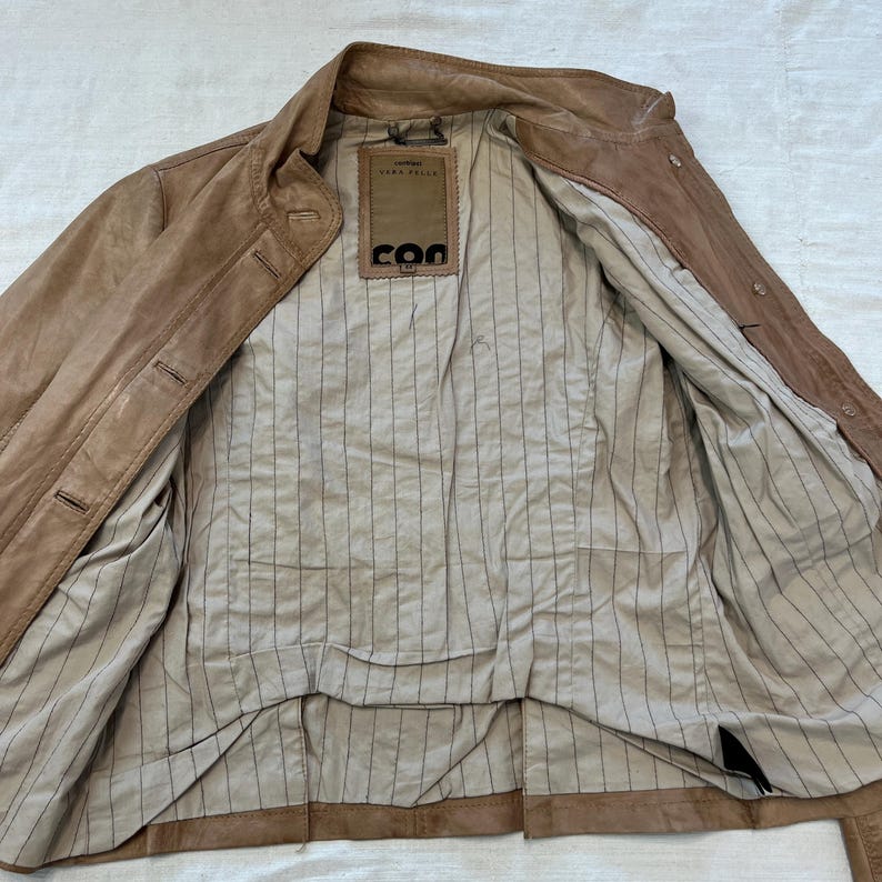 K&ouml;nnte beinhalten: Eine braune Lederjacke mit gestreiftem Futter. Die Jacke hat einen Knopfverschluss und einen Kragen. Das Etikett im Inneren der Jacke lautet "GINA PICCIOLI" und "COO".