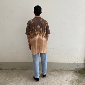 K&ouml;nnte beinhalten: Ein braunes Polo-Shirt mit Batikmuster und einem hellbraunen Kragen und &Auml;rmeln. Das Shirt hat ein einzigartiges gebleichtes Design, das von Dunkelbraun oben nach Hellbraun unten &uuml;bergeht. Die Person tr&auml;gt eine hellblaue Jeans und schwarze Turnschuhe.