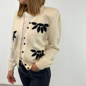 May include: A white cardigan with black floral embroidery. The cardigan has a button-up front and long sleeves.