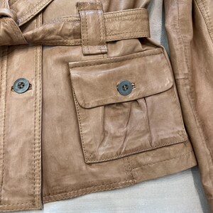 K&ouml;nnte beinhalten: Eine braune Lederjacke mit G&uuml;rtel und einer gro&szlig;en Patte auf der Vorderseite. Die Jacke hat einen Knopfverschluss und einen Knopf an der Tasche.