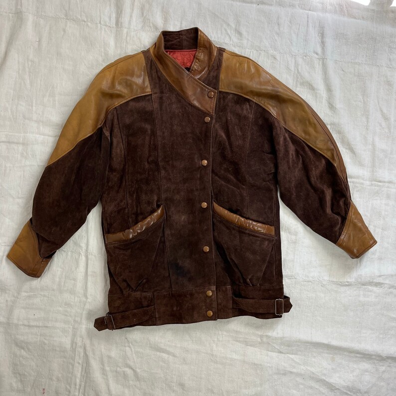 May include: A brown suede jacket with brown leather accents on the shoulders and sleeves. The jacket has a stand-up collar, a button closure, and two flap pockets.
