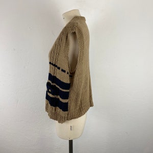 K&ouml;nnte beinhalten: Eine beige Strickweste mit marineblauen Streifen und einer Tasche. Die Weste wird auf einer Schaufensterpuppe vor wei&szlig;em Hintergrund pr&auml;sentiert. Die Weste hat eine gerippte Textur auf der Vorderseite und einen lockeren Strick an den Seiten und am R&uuml;cken.