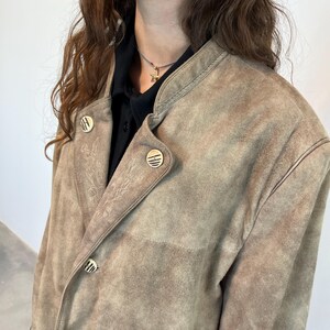 May include: A brown suede jacket with a stand-up collar and gold buttons. The jacket has a subtle embroidered design on the front.
