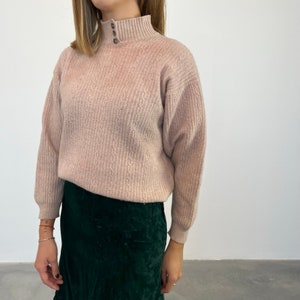 Può includere: Un maglione di maglia rosa chiaro con collo alto e chiusura a bottone. Il maglione ha un motivo testurizzato ed è indossato con una gonna verde scuro.