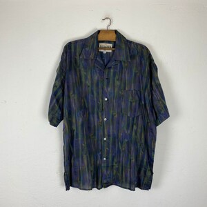 May include: A short-sleeved button-down shirt with a green, blue, and purple abstract print. The shirt has a collared neckline and a single chest pocket. The label on the shirt reads "Tavarnner".