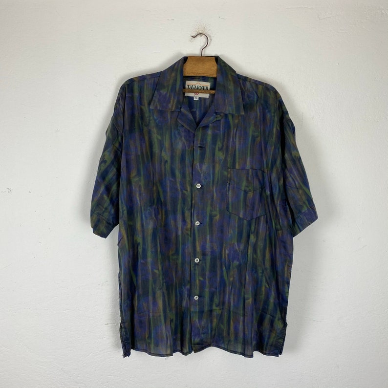 May include: A short-sleeved button-down shirt with a green, blue, and purple abstract print. The shirt has a collared neckline and a single chest pocket. The label on the shirt reads "Tavarnner".
