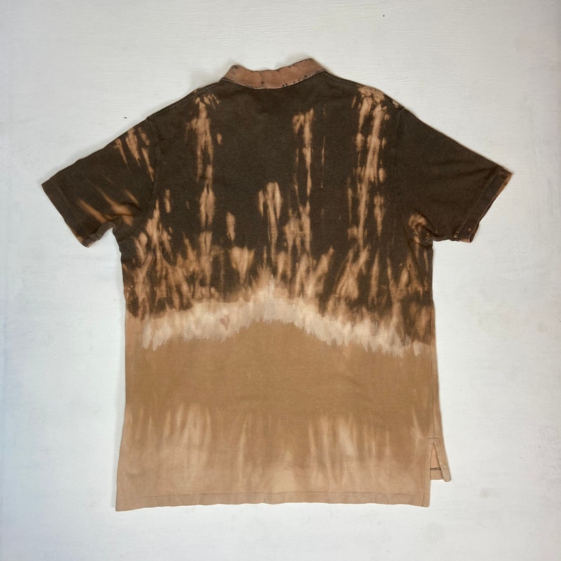 K&ouml;nnte beinhalten: Ein braunes Poloshirt mit einem einzigartigen Batik-Effekt. Der obere Teil ist dunkelbraun, der unten in ein helleres Braun &uuml;bergeht. Das Shirt hat einen Kragen und kurze &Auml;rmel mit einem geschlitzten Saum an den Seiten.