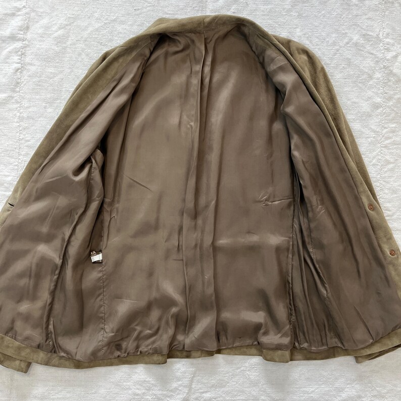 May include: A brown suede jacket with a button closure and a brown satin lining. The jacket is unbuttoned and laid flat.