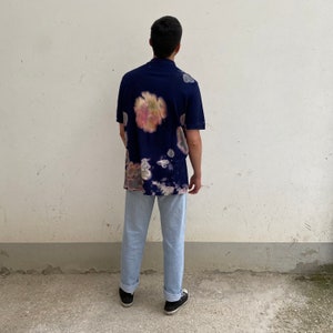 May include: A navy blue short-sleeved shirt with a tie-dye pattern in shades of pink, yellow, and white. The shirt is worn over light blue jeans.