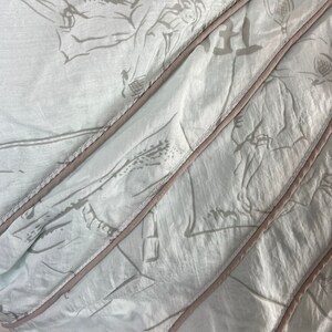 May include: Close-up of a light blue fabric with a subtle floral pattern in gray. The fabric is divided into diagonal sections with a light brown trim. The fabric appears to be soft and flowing, with visible folds and creases.