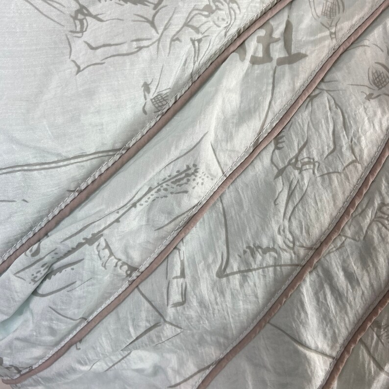 May include: Close-up of a light blue fabric with a subtle floral pattern in gray. The fabric is divided into diagonal sections with a light brown trim. The fabric appears to be soft and flowing, with visible folds and creases.