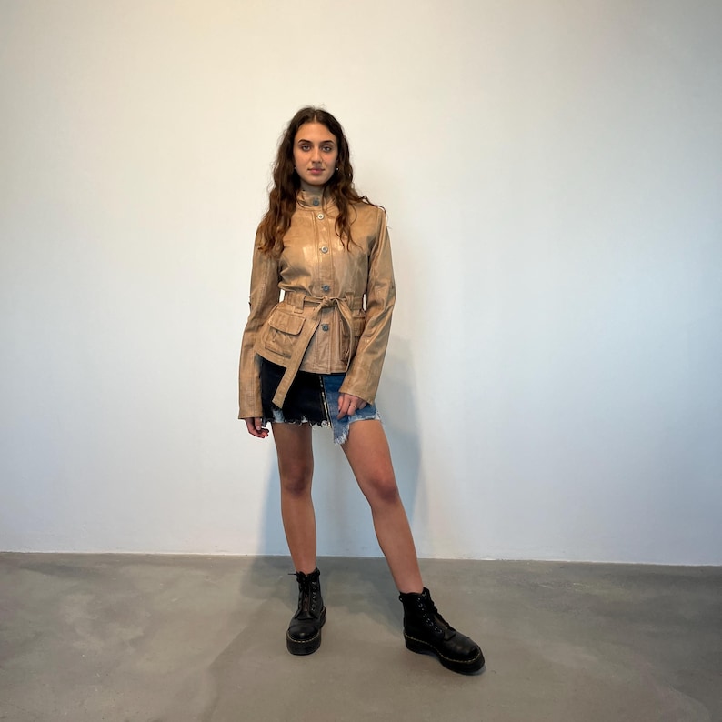 K&ouml;nnte beinhalten: Eine braune Lederjacke mit G&uuml;rtel und Taschen. Die Jacke hat einen Knopfverschluss und einen Stehkragen.