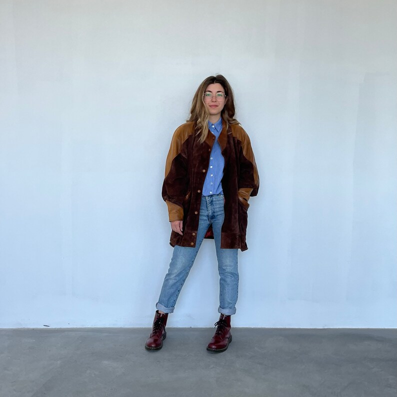 May include: A brown and tan suede jacket with a button closure. The jacket has a long, oversized fit and is worn over a light blue button-down shirt and blue jeans.